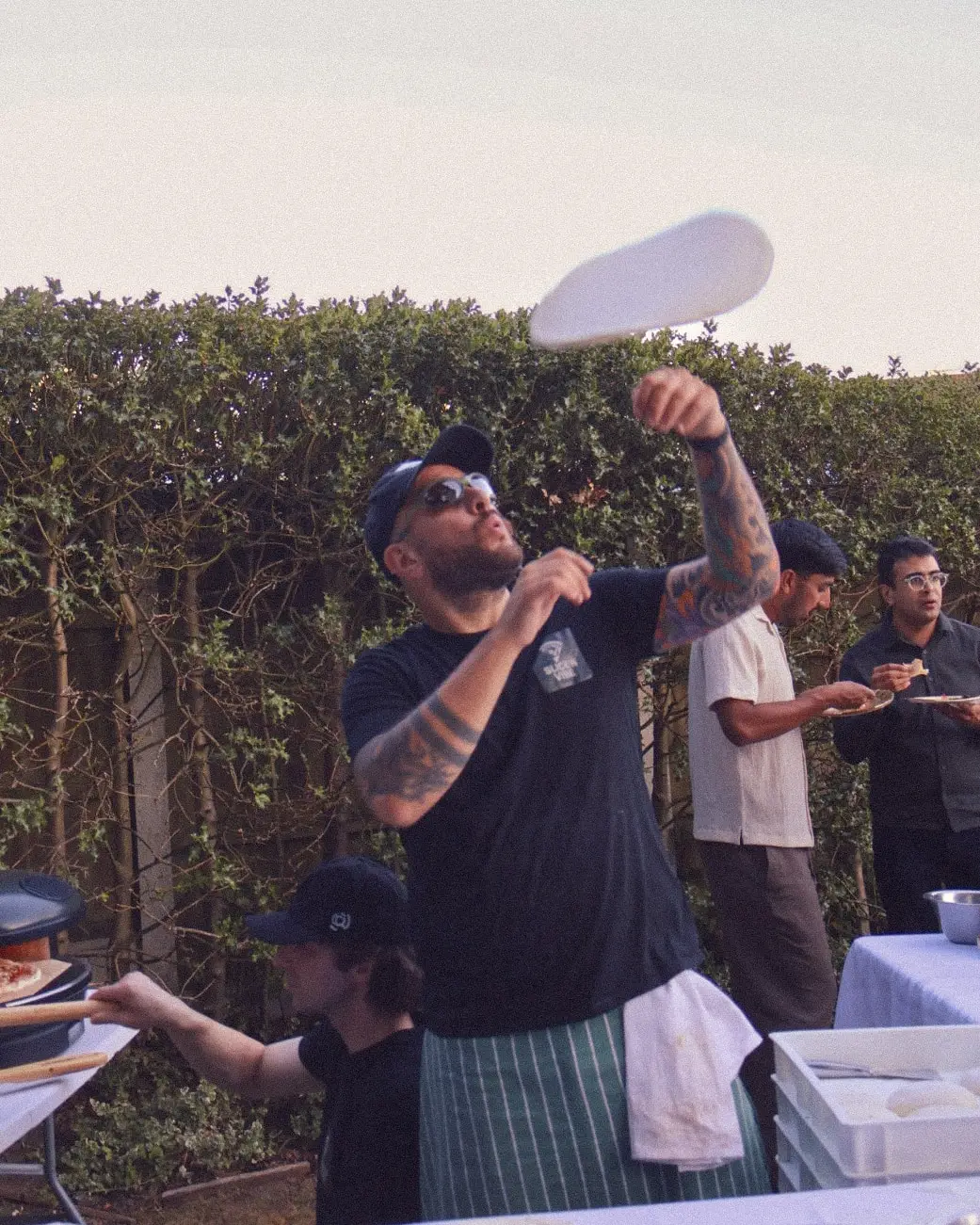 A pizzaiolo in a Slice 'N Vibe tee spinning a disc of dough above his head at dusk, guests eating behind him
